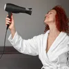 A woman with red curly hair in a white bathrobe is holding a black hairdryer and appears to be drying her hair. Produktbild-Vorschau