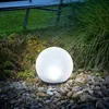 A glowing solar ball light sits in a garden bed surrounded by rocks and plants. Produktbild-Vorschau