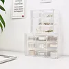 Two clear plastic drawer units are stacked on a white desk, one on top of the other. The top unit contains various jewelry items, while the bottom unit holds rolls of decorative tape. A framed quote is visible on the wall behind the units. Produktbild-Vorschau
