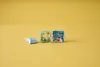 Two open toy boxes with a small doll in front of them, and a folded piece of paper to the left, on a yellow background. Anteprima dell’immagine prodotto