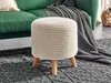 A beige knitted pouf with wooden legs sits on a dark gray patterned rug in front of a green sofa with a gray blanket draped over it. Náhled obrázku produktu