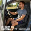 A man is sitting in the driver's seat of a car, holding the steering wheel. He is wearing a grey t-shirt and patterned shorts. A car seat cover is visible on the seat. Produktbild-Vorschau