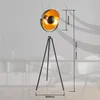 A black tripod floor lamp with a gold interior lampshade is shown against a blurred background of a room. Anteprima dell’immagine prodotto