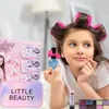 A young girl with hair rollers is holding a bottle of glitter nail polish and looking to the side. A makeup bag and other cosmetic items are in the foreground. Produktbild-Vorschau