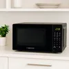 A black microwave oven is placed on a white countertop, with a small potted plant to its left and stacked plates on a shelf above. Anteprima dell’immagine prodotto