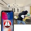 A security camera is shown mounted on a ceiling in a living room, while a person in a balaclava steals a laptop. A smartphone screen displays a security alert. Anteprima dell’immagine prodotto