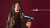 A woman holds a hairdryer in front of her face against a dark red background. Náhled obrázku produktu