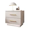 A white bedside table with two drawers and gold handles, a white lamp, and two perfume bottles on top. Produktbild-Vorschau