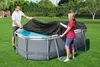 Two people are putting a black cover over an oval above-ground swimming pool in a grassy backyard. Podgląd zdjęcia produktu