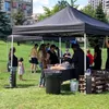 People gathered around a table under a black canopy in a park, with food items on display. Produktbild-Vorschau