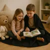 Two children are sitting on the floor, sharing a blanket and reading a book. Produktbild-Vorschau