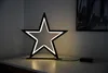 A black star-shaped LED lamp with warm white lights sits on a white surface, with a dark framed picture on the wall behind it. Produktbild-Vorschau