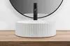 A white ceramic vessel sink with a fluted texture sits on a wooden countertop, with a black faucet and a round mirror in the background. Podgląd zdjęcia produktu