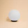 A single white globe light with a black base on a plain peach background. Produktbild-Vorschau