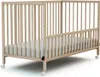 A light-colored wooden baby crib with a mattress inside, shown from a side-diagonal angle on a white background. Anteprima dell’immagine prodotto