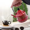 A person is pouring hot water from a red, decorative kettle into a small brown teapot. Voir les images du produit