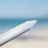 A close-up view of the tip of a silver umbrella pole with a white cap, set against a blurred background of the ocean and beach. Produktbild-Vorschau