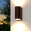 An outdoor wall light fixture is mounted on a textured white wall, with a tree and sunset visible in the background. Produktbild-Vorschau