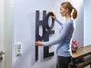 A woman is hanging a large metal 'HOME' decoration on a textured white wall. Anteprima dell’immagine prodotto