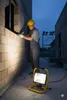 A construction worker is working on a brick wall at night, illuminated by a portable halogen lamp. Produktbild-Vorschau