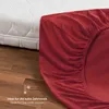 A red fitted sheet is shown draped over a mattress, with a portion of the mattress visible. The scene is set on a wooden floor. Voir les images du produit