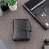 A black leather wallet is placed on a gray surface, with some small potted plants and a notebook with a card and pen in the background. Produktbild-Vorschau