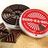A tin of Scho-Ka-Kola chocolate with some pieces of chocolate in front of it. Produktbild-Vorschau