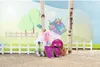 A doll's equestrian outfit and accessories are displayed outdoors, with a backdrop of trees and a fence. Produktbild-Vorschau