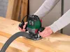 A person is using a PARKSIDE® Oberfräse (router) on a wooden surface, with a vacuum hose attached for dust collection. Produktbild-Vorschau