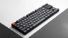 A black mechanical keyboard with an orange escape key is placed diagonally on a surface divided into white and dark gray sections. Produktbild-Vorschau