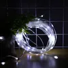 A coiled string of white LED lights is placed next to a green plant on a wooden surface, with a grid-like background. Produktbild-Vorschau