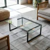 A modern living room with a glass coffee table on a rug, near a sofa and a woven pouf. Produktbild-Vorschau
