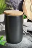 A black ceramic storage jar with a wooden lid sits on a dark surface dusted with powdered sugar. Other kitchen items and plants are visible in the background. Produktbild-Vorschau