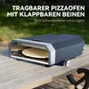 A portable pizza oven with folding legs sits on a wooden table outdoors, with greenery in the background. Produktbild-Vorschau