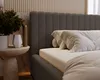 A close-up view of a dark gray bouclé fabric bed with white pillows and bedding, next to a small side table with a plant and vase. Náhľad obrázku produktu
