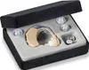 A beige hearing aid is displayed in a black case with a grey foam insert. The case also holds three clear ear tips and two silver batteries. Produktbild-Vorschau