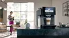 A woman is using her phone in a modern kitchen while a Siemens coffee machine is in the foreground. Produktbild-Vorschau