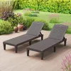 Two brown plastic sun loungers are placed on a wooden deck in a garden setting, with lush green bushes and flowering plants in the background. Podgląd zdjęcia produktu