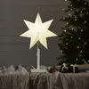 A white star-shaped lamp sits on a table covered with a beige cloth, with a decorated Christmas tree in the background. Podgląd zdjęcia produktu