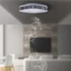 A smoke detector on a ceiling with smoke rising from it, in a living room setting. Podgląd zdjęcia produktu