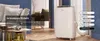 A white portable air conditioner is shown in a living room setting, with text overlays highlighting its features. Produktbild-Vorschau