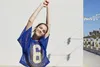 A young woman stretches her arms up while wearing a blue mesh jersey with the number 6 on it, standing outdoors with a white wall behind her and a beach scene in the background. Produktbild-Vorschau