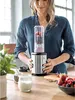 A woman is holding a mini smoothie maker filled with berries and a pink smoothie in a kitchen setting. Produktbild-Vorschau