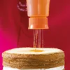 A person is using an orange squeeze bottle to pour liquid onto a cake. Podgląd zdjęcia produktu