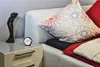A close-up view of a bedside table next to a bed, featuring a small alarm clock and a decorative figurine. Produktbild-Vorschau