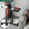 A practical end table with a marble-like top is shown next to a sofa, with a drink and a clock on it. A cat is lying underneath the table. The base of the table with wheels is also visible. Produktbild-Vorschau