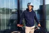 A man wearing a helmet and hoodie stands with his bicycle in front of a modern building with reflective windows. Produktbild-Vorschau