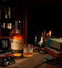 A bottle of Glen Garioch whisky and a glass of whisky sit on a wooden table in front of a fireplace. Books and a camera are also on the table. Produktbild-Vorschau