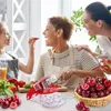 Three women of different ages are in a kitchen preparing food. A cherry pitter is in the foreground, and a basket of cherries is to the right. Náhled obrázku produktu