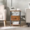 A small wooden side table with a drawer and shelves, placed between two armchairs in a living room setting. Produktbild-Vorschau